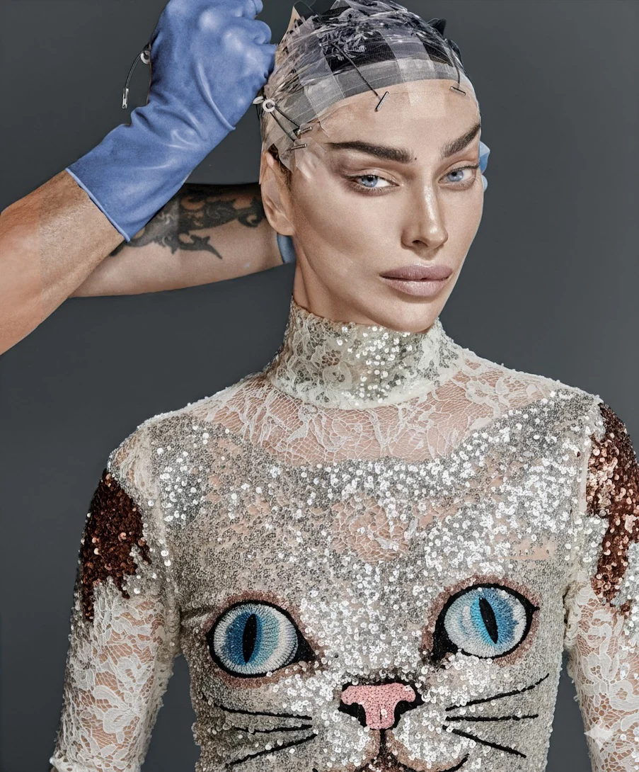 Vogue beauty portrait of a model in a plaid cap and a sequined top with an embroidered cat face, a blue-gloved hand at her head.