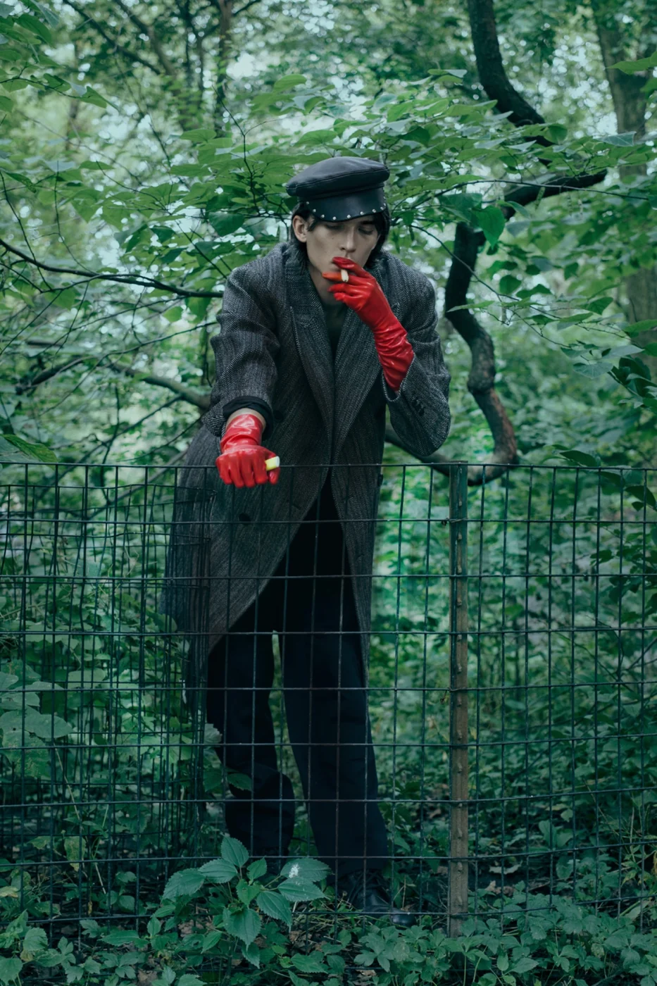 Dsquared2 image in a park: a man in a long coat and cap, wearing red gloves, standing at a wire fence.